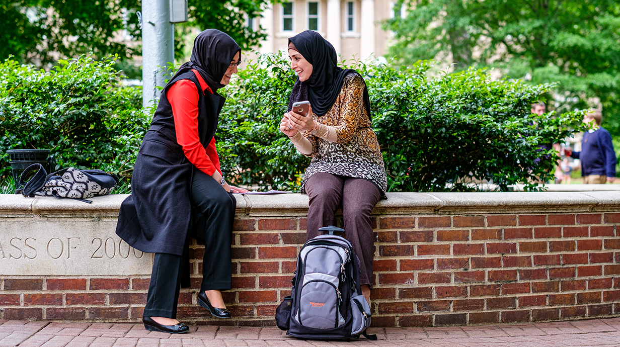 Two women sit smiling outside on the Ole Miss campus, one showing the other something on her iPhone.
