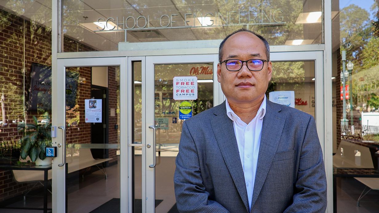 Dr. Xiaopeng Zhao stands in front of Carrier Hall on the Ole Miss campus with School of Engineering showing in the window.