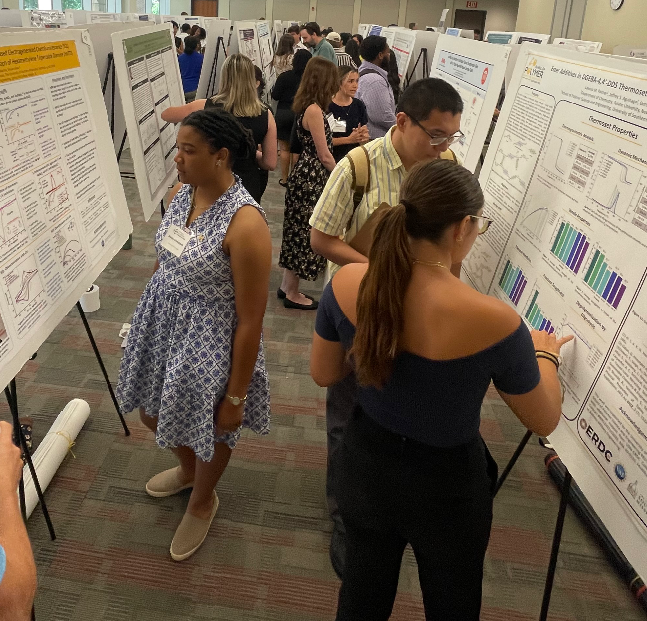 Students stand in front of large posterboards explaining research projects to others