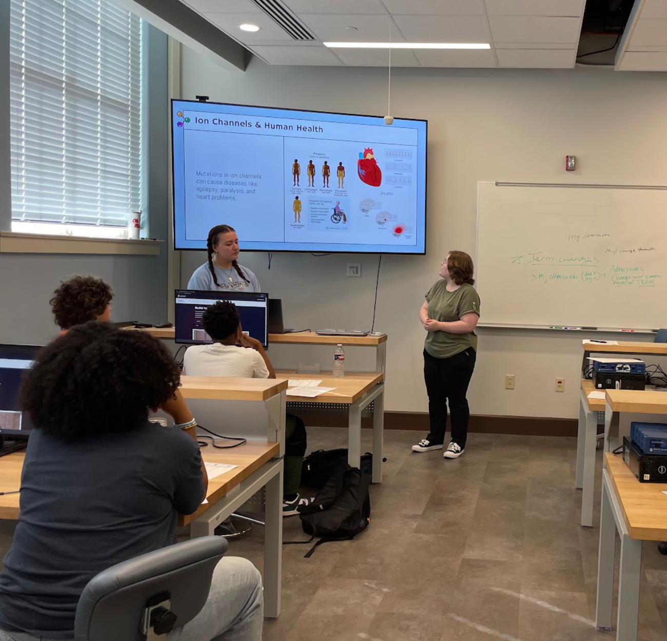 Two students stand at the front of a classroom where a powerpoint presentation displays a slide titled "Ion Channels & Human Health"