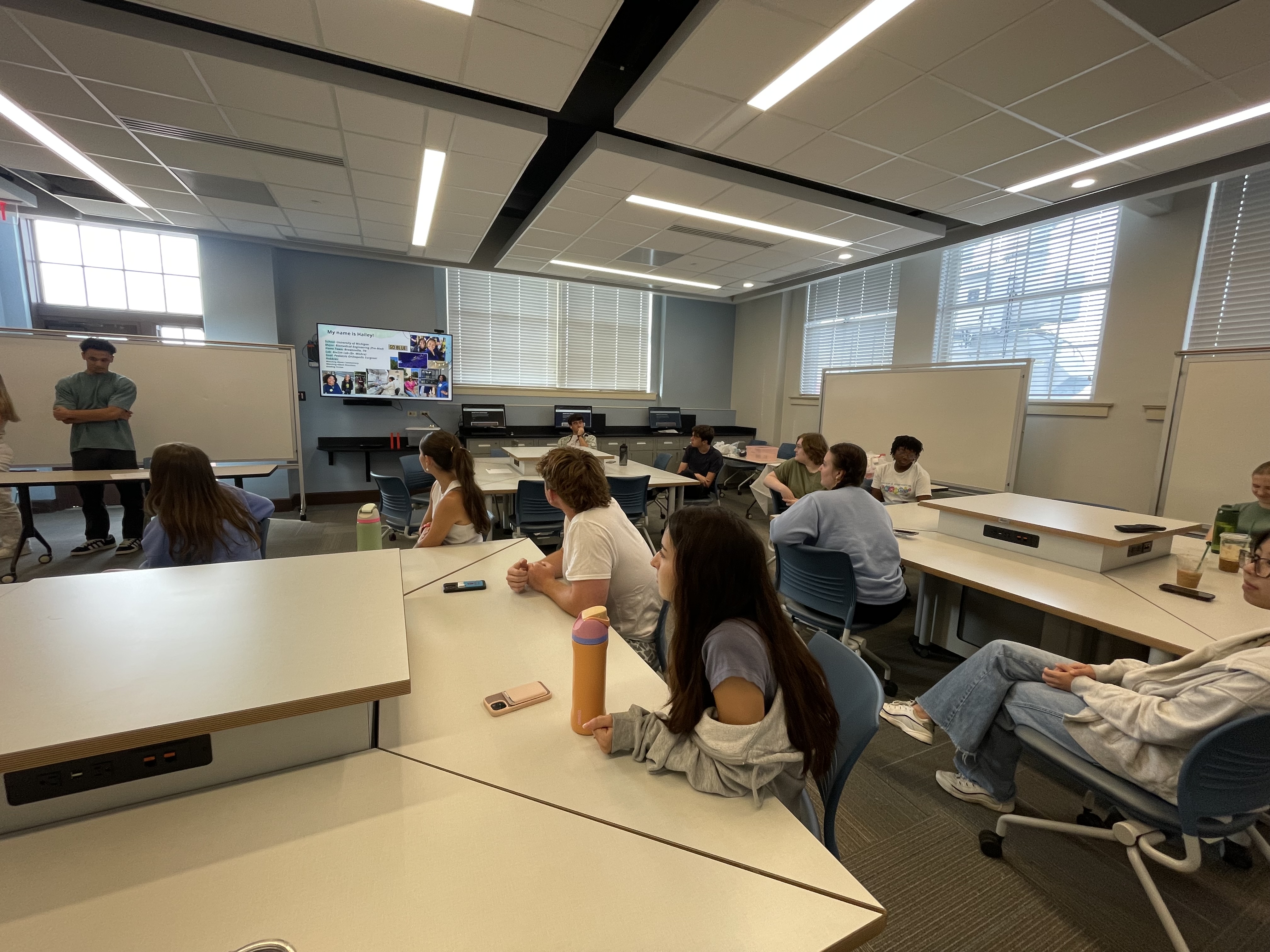 High school students sit listening to undergraduate students at the front of a classroom explain a concept