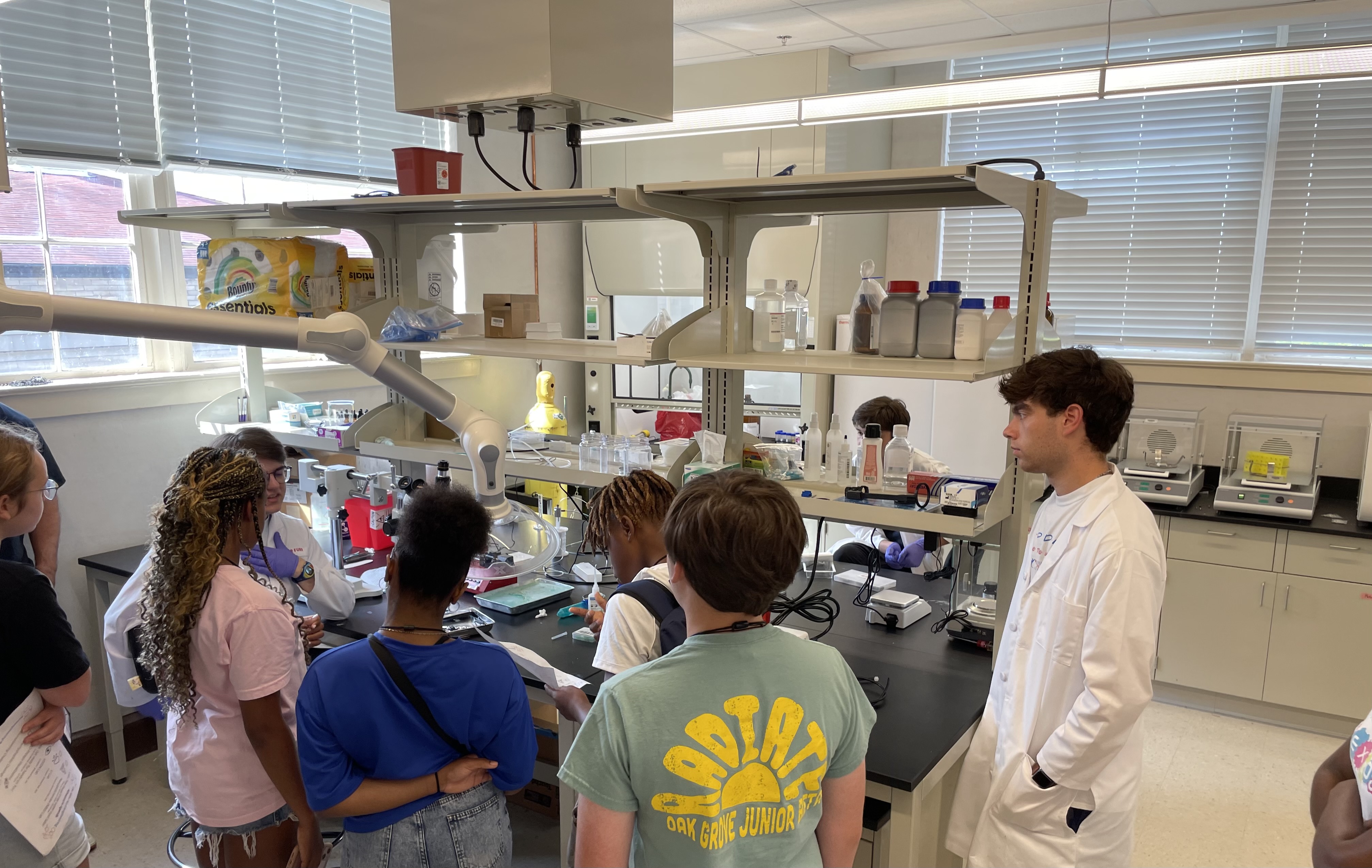 Two undergraduate students wearing lab gear teach a group of middle school students about concepts in the lab.