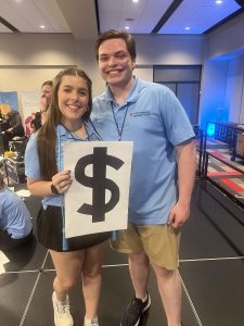A smiling young woman and man, both in RebelTHON blue polos and with colorful face paint, proudly display a large dollar sign signifying the successful fundraising efforts at the RebelTHON event.