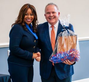 A woman and a man holding a gift basket wrapped in clear plastic with red and blue decorative fillers. They are both smiling and dressed in business attire. The woman is wearing a navy blue suit with a blue scarf, and the man is in a blue suit with a light blue shirt and patterned tie. The setting appears to be the same indoor room as the previous photo, with a simple wall and minimal decor.