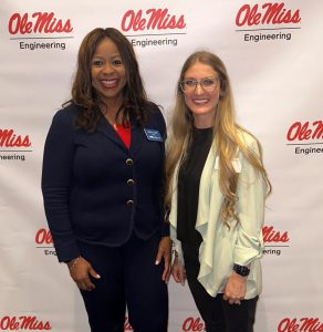 Two women smiling for the camera in front of a banner that reads "Ole Miss Engineering" with the university's logos. The woman on the left is wearing a navy blazer and trousers with a blue scarf, while the woman on the right is in a light-colored top and dark trousers. They appear to be at an indoor event.