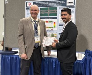 Gaurav Aryal, a graduate student in a black suit, smiling and holding an award certificate, stands next to a man in a beige suit in front of a research poster about Restricted Crossing U-Turn (RCUT) intersections. The poster contains charts and text about the study's findings."